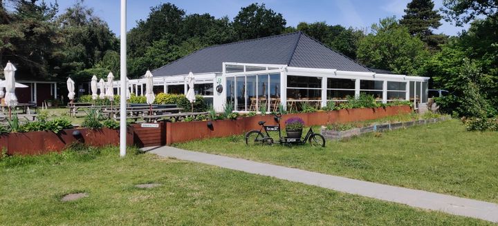 Udendørsbillede af den hvide Restaurant Kronborg på Egholm med grønne arealer, udendørsborde og en ladcykel i forgrunden.