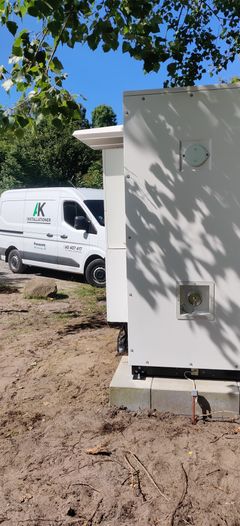 Nærbillede af et stort, hvidt kommercielt batterianlæg fra Solis udendørs på en grusplads. I baggrunden holder en hvid AK Installationer firmabil under grønne træer.