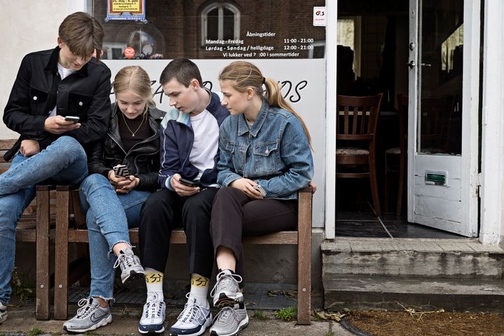 Fire unge sidder på en bænk foran en butiksfacade og kigger på deres telefoner.