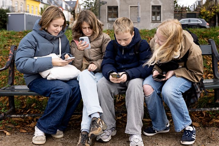 Fire børn sidder på en bænk og bruger deres smartphones.