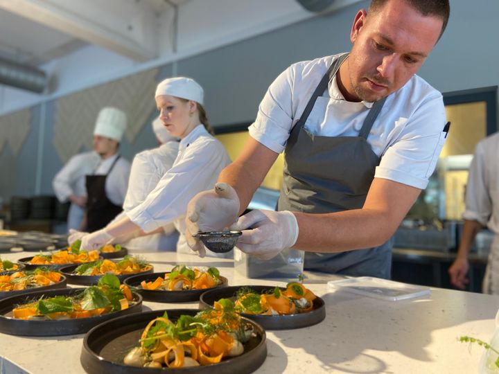 En kok anretter mad på en tallerken i et køkken, mens andre kokke arbejder omkring ham.