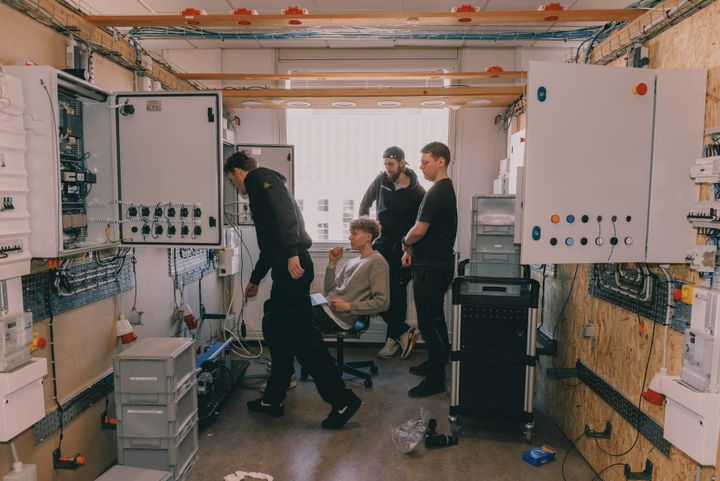 Fire personer arbejder sammen i et teknisk lokale med kontrolpaneler og udstyr.