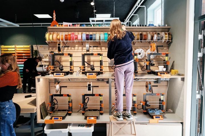 En person står på en stige foran en væg med farverige trådruller og 3D-printere.