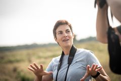 En person i en naturkulisse, iført en grå T-shirt og en kamerastrop om halsen, gestikulerer med hænderne.