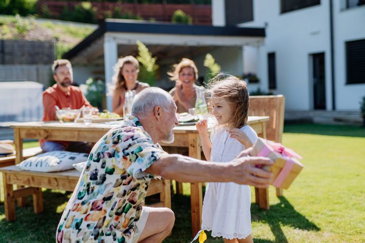 En ældre mand giver en fødselsdagsgave til en lille pige i en have. Familie sidder ved et bord i baggrunden.