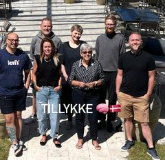En gruppe mennesker står sammen og smiler udendørs. Tekst: "TILLYKKE" med emoji af en hjerne og det danske flag foran dem.