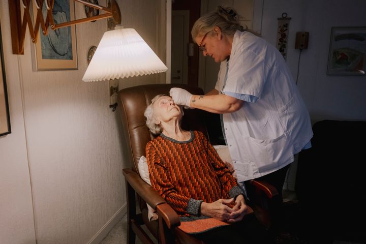 En ældre kvinde sidder i en stol, mens en sundhedsmedarbejder undersøger hende under en lampe.