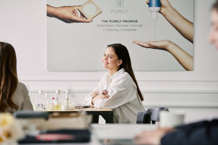Kvinde sidder ved et bord i et mødelokale med hænder foldet foran sig og smiler. I baggrunden er der en reklameplakat med hudplejeprodukter.