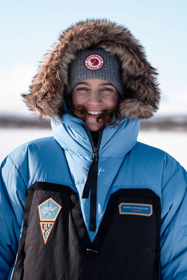 En person iført en blå jakke med pelskrave og en grå hue, smiler udendørs i et vinterlandskab.