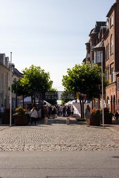 Bygade med brostensbelægning og træer langs fortovet. Mennesker går mellem stande og butikker.