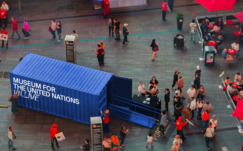 Mennesker står omkring en portal: en stor blå shippingcontainer med skriften Museum for The United Nations UN Live på siden