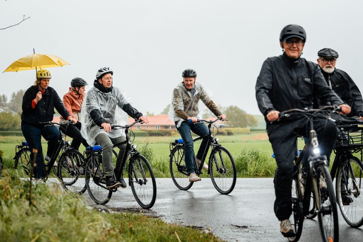 En gruppe mennesker cykler i regnvejr. Nogle bærer regnjakker, andre regnslag og en person cykler med en udslået gul paraply