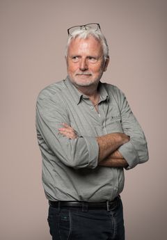 Jeg ved ikke, hvem personen på billedet er, men her er et forslag til alt-tekst: En ældre mand med gråt hår og skæg står med arme over kors. Han bærer en grå skjorte og briller på hovedet.