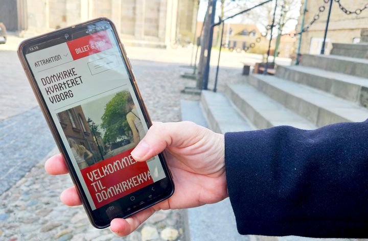 En hånd holder en smartphone foran trapperne ved Viborg Domkirke. På telefonens skærm ses en hjemmeside for Domkirkekvarteret.dk med tekst om billetter og velkomst.