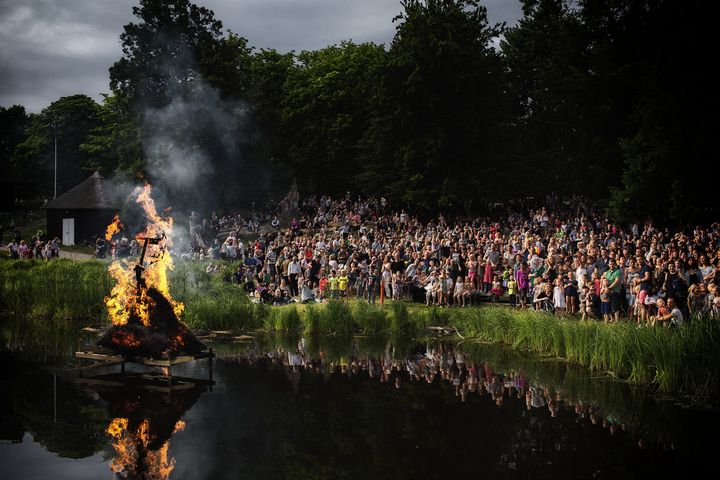 Folk samlet omkring et Skt. Hans bål ved en sø.