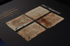 Fire gamle, brunfarvede pergamentmanuskriptsider spredt ud på et bord med en farveskala og tre små træpinde ved siden af.