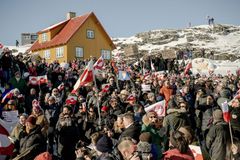 Stor folkemængde med grønlandske flag foran et gult hus i sneklædt fjeldlandskab.