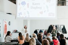 **Alt text:** En Det Kgl Biblioteks direktør Bente Skovgaard Kristensen taler til en gruppe skoleelever foran et stort skærmbillede med teksten "Kære Dagbog". Eleverne sidder ved borde og lytter.