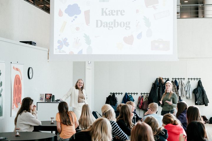 **Alt text:** En Det Kgl Biblioteks direktør Bente Skovgaard Kristensen taler til en gruppe skoleelever foran et stort skærmbillede med teksten "Kære Dagbog". Eleverne sidder ved borde og lytter.