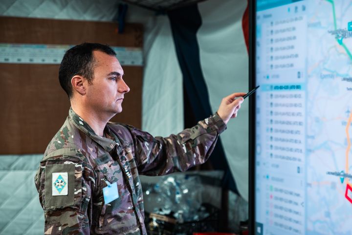 French Military personnel in uniform pointing at a large digital display screen with maps and data.