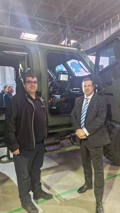 Two people standing in front of a military vehicle in an indoor setting.