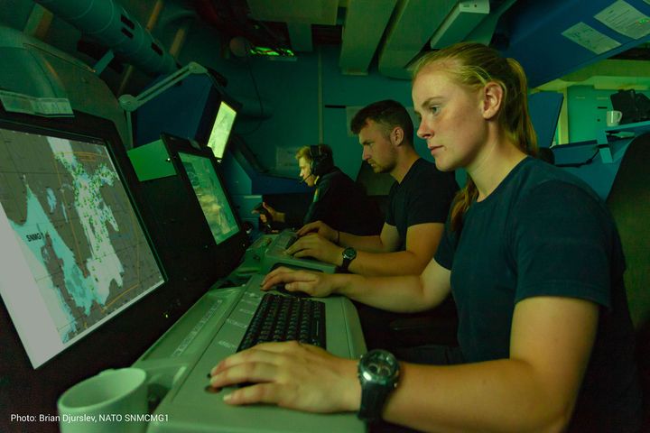 Three people work intently at computer stations in a dimly lit room with maps displayed on screens.