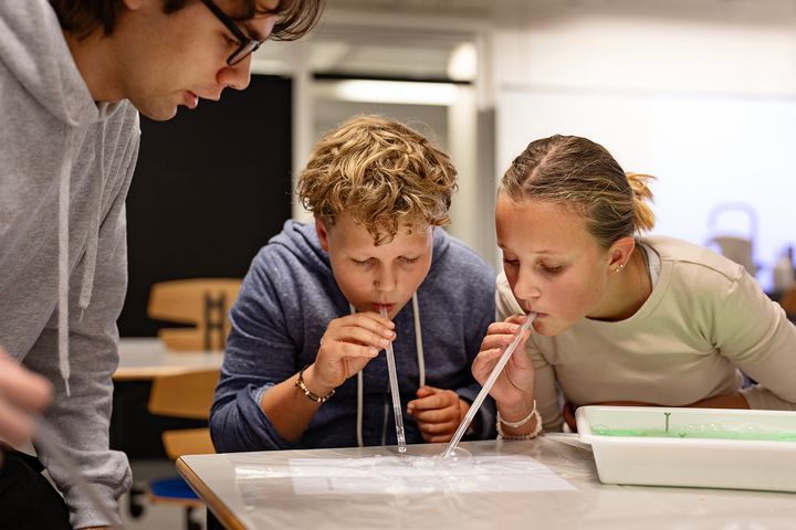 To børn puster til en bakke med sæbevand ved hjælp af sugerør, mens en ung dreng står og kigger på dem.