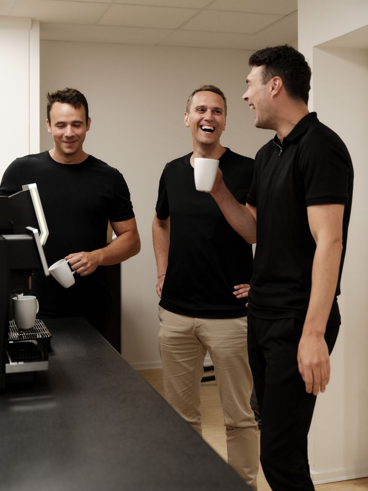 Tre personer står ved en kaffemaskine og griner sammen.