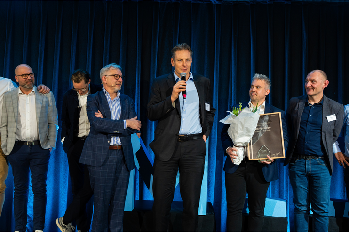 Rynkebys direktør Mark Ole Juel Hemmingsen står på scenen til MLDK Årstræf og tager imod prisen for "Årets Mærkevareleverandør: Socialt Engagement". Ved siden af står MLDK's direktør Leif Nielsen og i baggrunden tidligere ryttere, der har cyklet for Team Rynkeby.