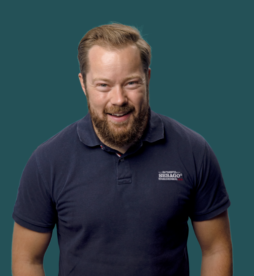 Jeg ved ikke, hvem personen er, men billedet viser en mand i en mørkeblå polo mod en ensfarvet baggrund.