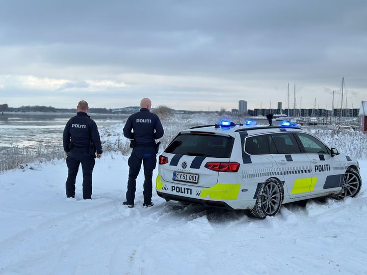 To%20betjente%20og%20en%20politibil%20med%20bl%E5%20blink%20st%E5r%20p%E5%20en%20sned%E6kket%20vej%20ved%20vandet.