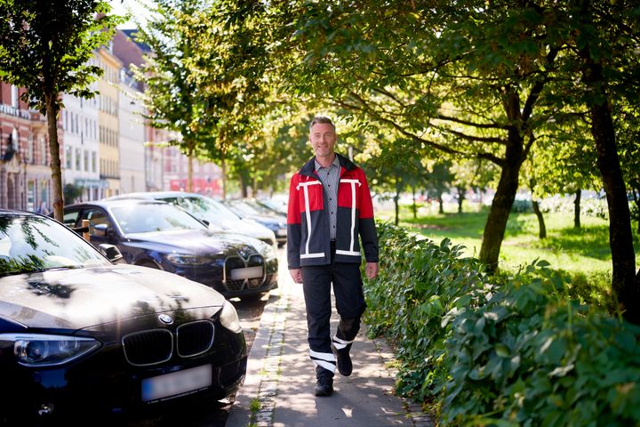 Person iført Q-Park-uniform går på fortov i solskinsvejr, med parkerede biler langs vejen.