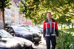 Person iført Q-Park-uniform går på fortov i solskinsvejr, med parkerede biler langs vejen.