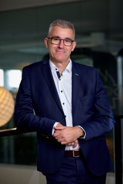 En mand i et jakkesæt står smilende med hænderne foldet foran sig.