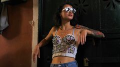 Here's a short alt text suggestion:
"A person with a tattooed arm wearing jeweled sunglasses and a bejeweled top, posing casually."