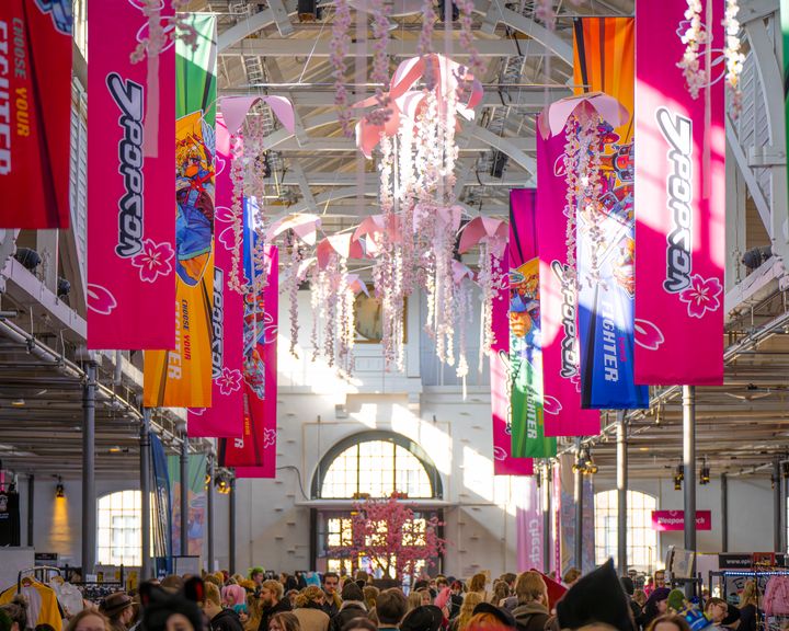 Farverige bannere med J-Popcons navn og maskotter hænger fra loftet i Øksnehallen sammen med store lyserøde blomster, mens mennesker strømmer ind.