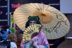 En pige i grøn paryk, lyserød kjole og med en stor paraply holder en gul vifte op foran sit ansigt.