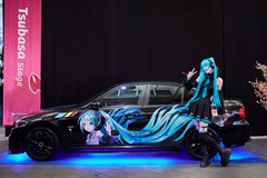 En sort bil med en animepige med blåt hår står med blåt lys under sig. Ved siden af står en pige i et kostume, der matcher pigen på bilen.