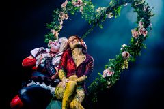 En kvinde i et klovnelignende kostume med hvid ansigtsmaling og blond paryk læner sig op ad en grøn kvinde dækket i blade med et rødt kostume og rødt hår, begge smilende. Bag dem er en kæmpe hjerte dækket i blomster og grønt.