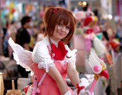 Pige i brun paryk, lyserød kjole, hvide vinger og med stav i hånden poserer for kamera.