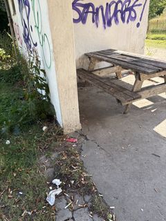 BIlledet viser en bunke cigaretskodder på jorden lige foran et overdækket område, med et borde-bænkesæt, som er fyldt med grafitti