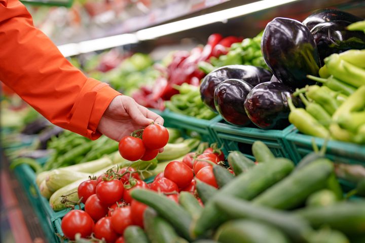 En person i orange jakke vælger tomater i en grøntsagsafdeling med agurker, auberginer og peberfrugter.