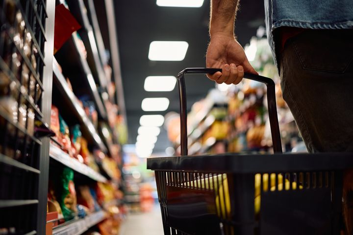 En person bærer en indkøbskurv i en supermarkedsgang fyldt med varer.