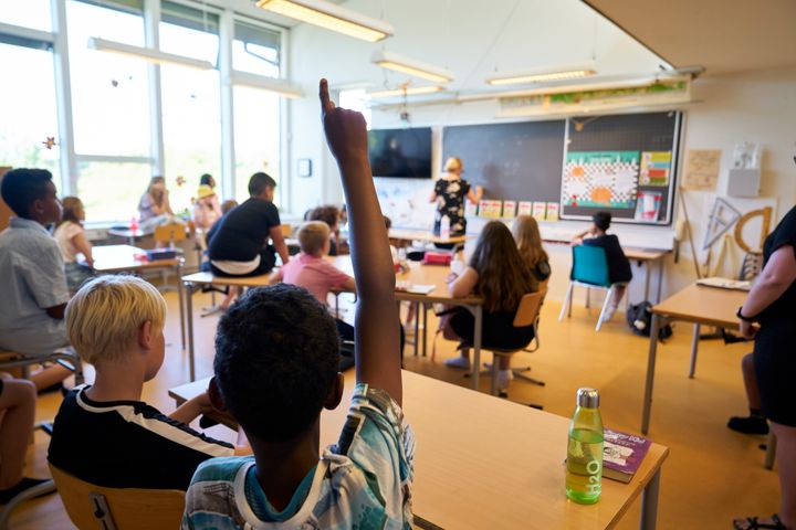 Undervisning i et klasseværelse på mellemtrinnet hvor man ser eleverne bagfra og læreren ved tavlen. Et barn rækker hånden op.