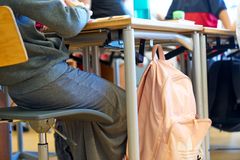 Skoletaske der hænger på et skolebord og en elev sidder på stolen ved bordet med ryggen til