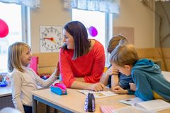 En smilende kvindelig lærer sidder ved et skolebord med børn rundt om og hun har øjenkontakt med et af børnene