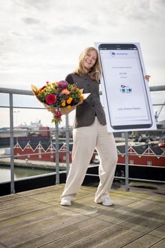 En tydelig glad Julie Winther Nielsen står på toppen af et tag med udsigt over Frederikshavn Havn i baggrunden. Hun holder blomster i den ene hånd og en mobilepay-check på kr. 30.000 i den anden. Det er det synlige bevis på, at hun har vundet prisen.