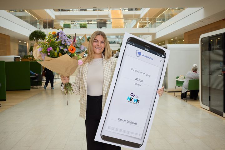 En glad Yasmin Lindharth holder blomster og en MobilePay-check op for at vise, at hun har modtaget årets fagprøvepris 2026.
