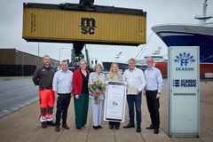 Syv personer deltager i fejrningen af vinderen. De står foran en container og vinderen holder blomster og check som symbol på prismodtagelsen.