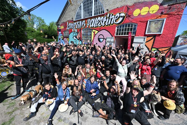 En stor gruppe mennesker klædt i sort jubler og vinker med armene foran en rød bygning med graffiti, hvor der bl